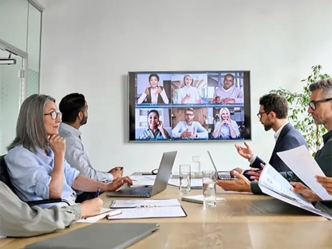 People in a conference room having a meeting with colleagues on a video call displayed on a large screen.