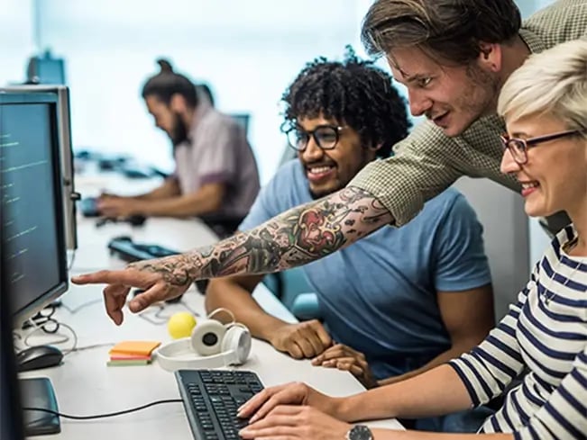 Person with tattoos points at a computer screen as a colleague types on a keyboard in an office.