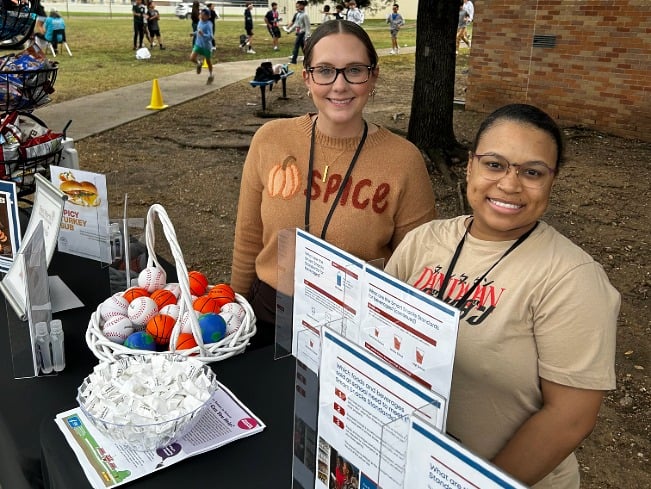 Two dietitians stand outside a school behind a table displaying nutrition information and resources. 