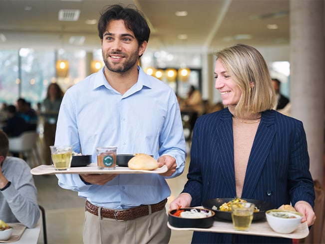 Two people carrying lunch trays with plates and drinks while walking through a bright cafeteria.
