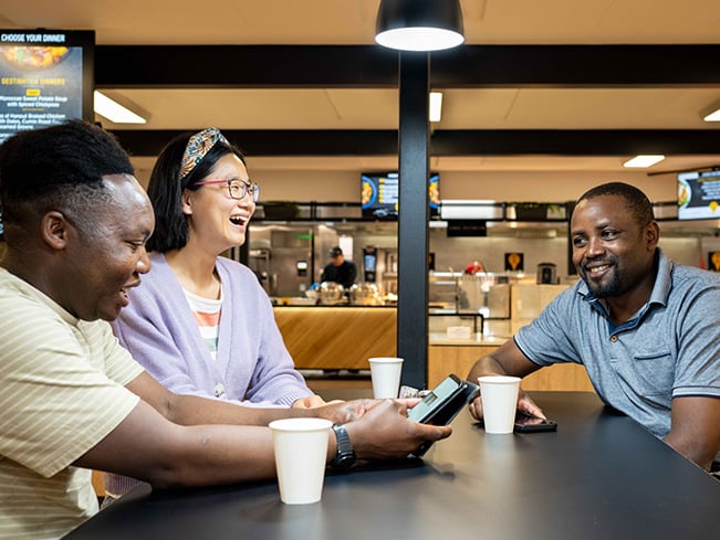 Three people seated at a cafeteria table with paper cups, one holding a smartphone, food counters in the background.