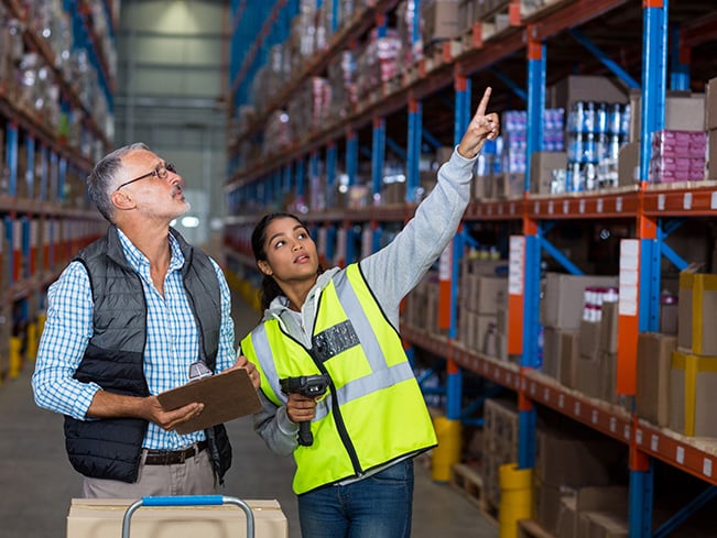 Two warehouse workers review inventory, one pointing at shelves stacked with boxes and goods.
