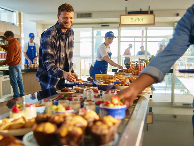 People selecting pastries and desserts from a buffet counter in a bright cafeteria with staff working behind the counter.
