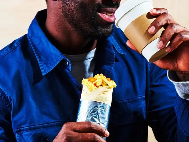 A person in a blue shirt holding a foil-wrapped burrito in one hand and a coffee cup in the other, about to sip.