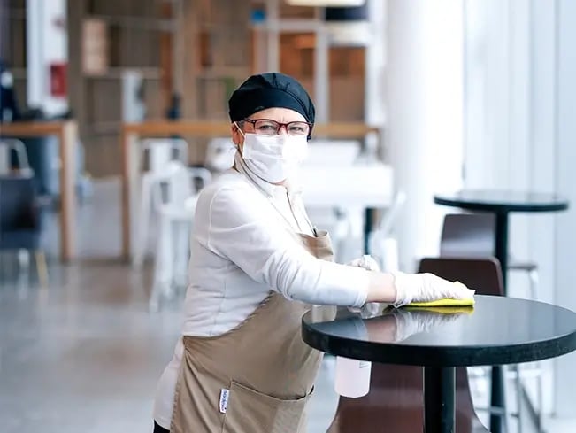 Cafe worker in beige apron and gloves wipes a round black table with a yellow cloth.