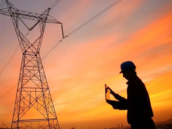 Silhouette of person with tablet by power line tower at sunset.