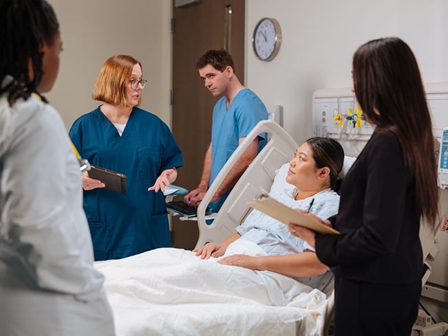 Care team discusses treatment at a patient’s bedside in a hospital room with charts and medical equipment.