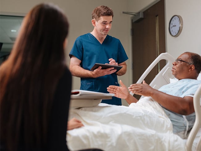 Healthcare professional reviewing information on a tablet while speaking with a patient in a hospital room.