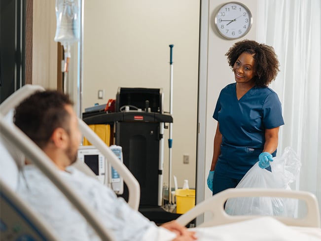 Healthcare worker collecting waste in a patient room while speaking with a patient beside a hospital bed.