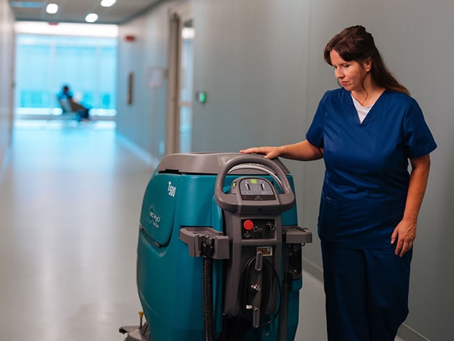 Healthcare worker operating an automated floor-cleaning machine in a hospital hallway.