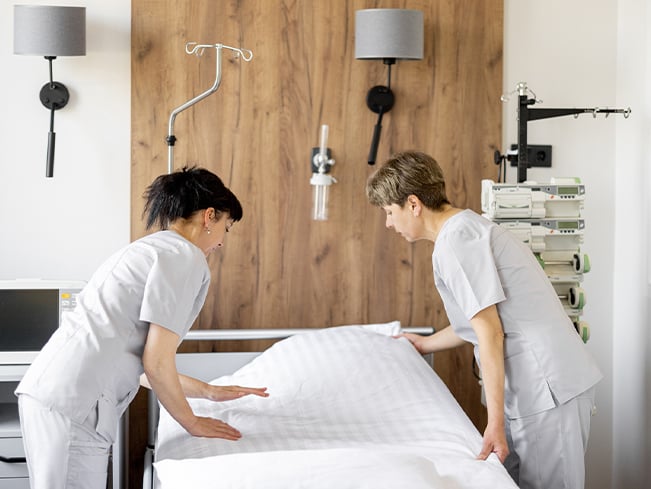 Healthcare staff making a hospital bed in a patient room, with medical equipment and bedside fixtures visible, highlighting cleanliness, room preparation, patient safety, and healthcare support services.