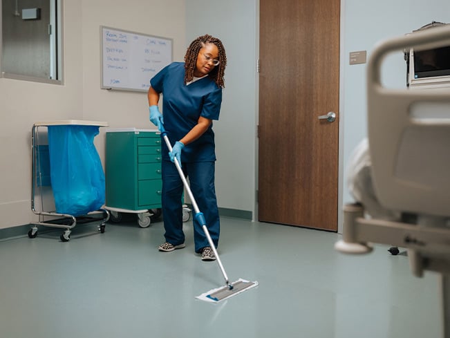 Healthcare environmental services staff member mopping the floor in a patient room, highlighting hospital cleanliness, infection prevention, sanitation protocols, and safe care environments.