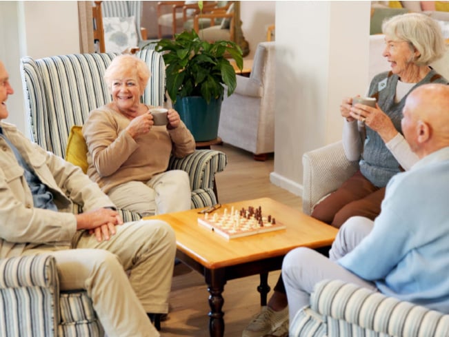 Older adults gathered in a comfortable lounge area, enjoying conversation over coffee around a table with a chessboard, highlighting social engagement, community living, and wellness-focused shared spaces.
