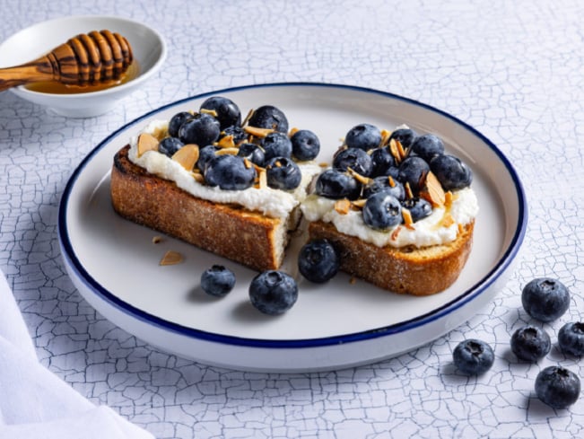 Blueberry toast topped with creamy spread and sliced almonds on artisan bread, served on a plate with fresh blueberries and honey, highlighting a nutritious breakfast and wholesome dining option.