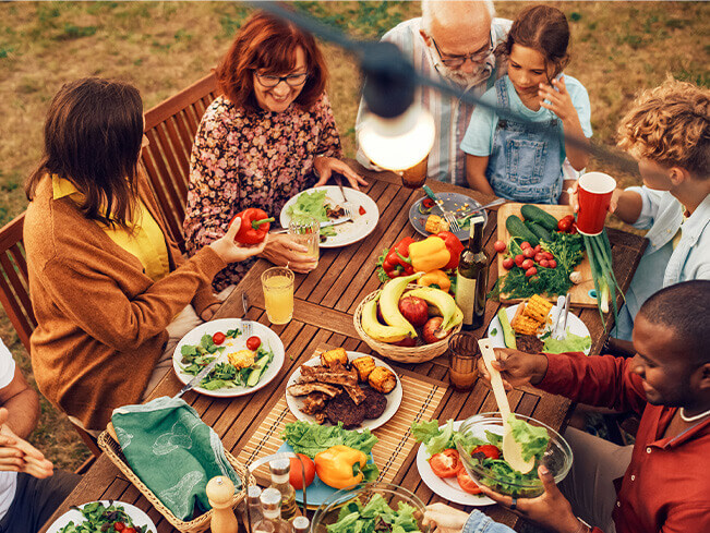 Multigenerational group gathered around an outdoor dining table with fresh vegetables, salads, and grilled foods, highlighting shared meals, social connection, healthy eating, and community-centered dining experiences.
