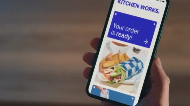 Close-up of a hand holding a smartphone displaying a food service app with a notification reading “Your order is ready” and an image of a wrap meal, set against a softly blurred background.