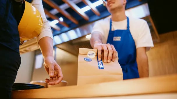Close-up of a takeout interaction at a counter, with a food service worker handing a small paper bag to a customer, highlighting contactless pickup in a modern dining setting.