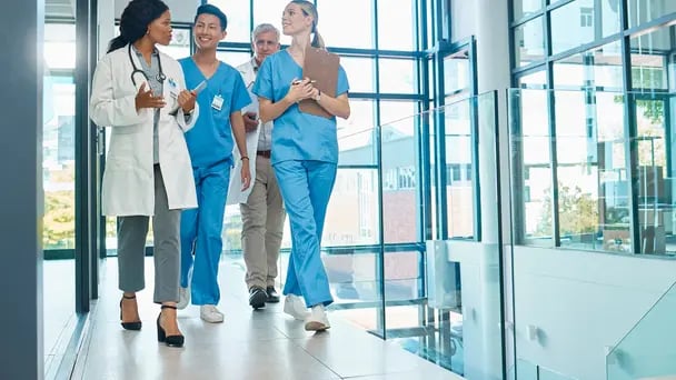 Group of healthcare professionals walking through a bright hospital corridor, wearing medical scrubs and a lab coat, holding a clipboard, and talking in a modern clinical setting with large windows.