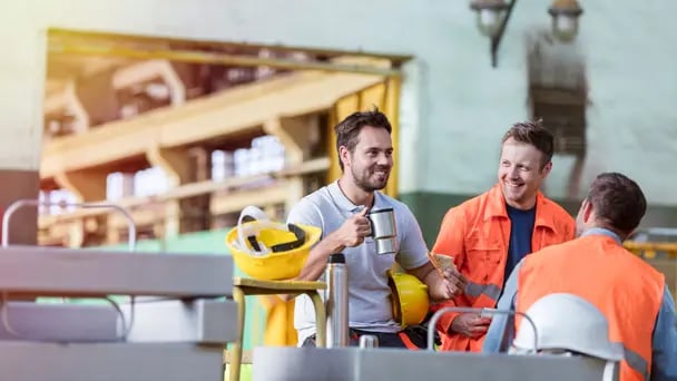 Three workers in a break area, wearing safety gear including helmets and high-visibility jackets, sitting together, holding coffee and chatting near industrial equipment.