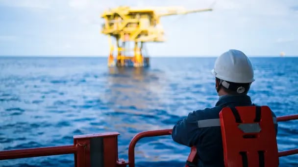 Worker wearing a helmet and safety gear standing on a vessel, looking toward an offshore platform over the ocean, with blue water and industrial structure in the distance.