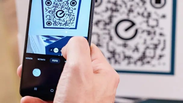 Close-up of a hand using a smartphone to scan a QR code displayed on a wall, with the camera interface visible and a large code pattern in the background.