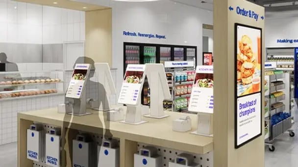 Self-service ordering area in a modern cafeteria with multiple touchscreen kiosks on a counter, beverage dispensers below, and a large digital menu display showing food options on the wall.