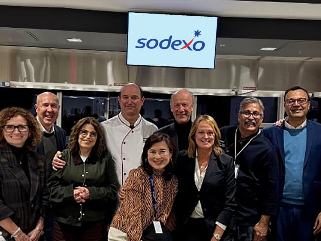Group of professionals standing together in front of a Sodexo sign inside a commercial kitchen or event space, posing for a group photo during a company gathering.
