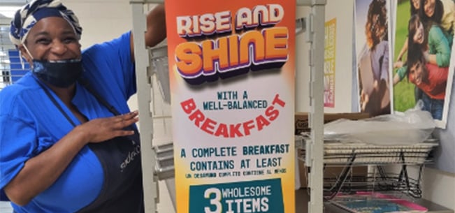 Food service staff member standing beside a breakfast cart featuring a Rise and Shine promotional sign with information about complete breakfast items, inside a school food service area.