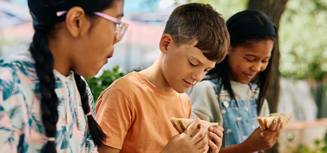 Three children sitting outdoors together holding sandwiches, looking at their food while eating in a shaded area with trees and greenery visible in the background.