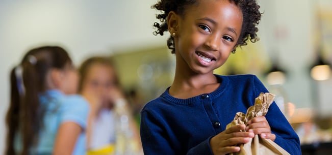 Child holding a crumpled paper bag indoors, with other children seated at tables in the softly focused background of a bright school cafeteria or classroom setting.