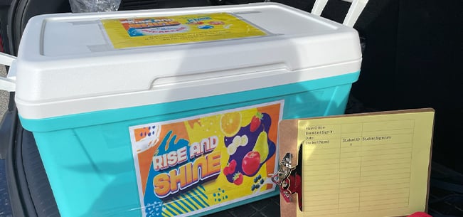 Blue insulated cooler labeled Rise and Shine placed in the back of a vehicle, next to a clipboard with a meal distribution sign in sheet and colorful fruit themed graphics.