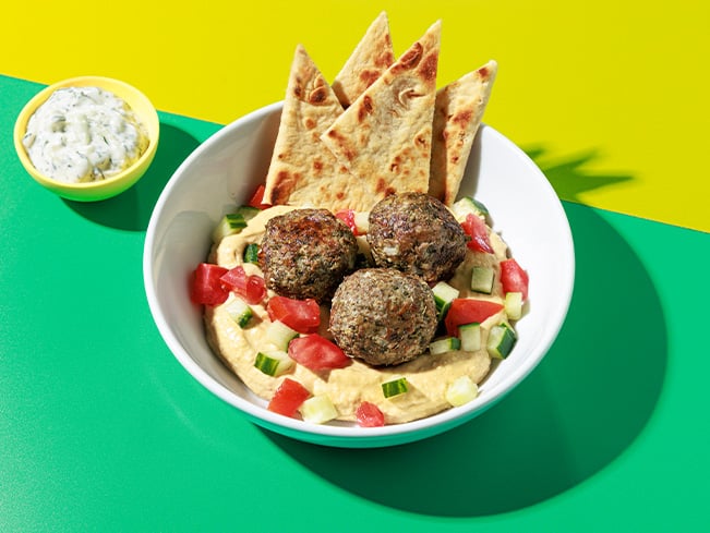 Bowl of hummus topped with falafel, diced cucumbers, and tomatoes, served with grilled pita wedges and a side of tzatziki dip, showcasing a fresh and nutritious food service menu option.