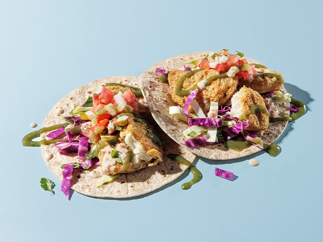 Two soft tortilla tacos topped with crispy breaded protein, shredded red cabbage, fresh salsa, sliced peppers, and green sauce, showcasing a vibrant and freshly prepared school dining meal option.
