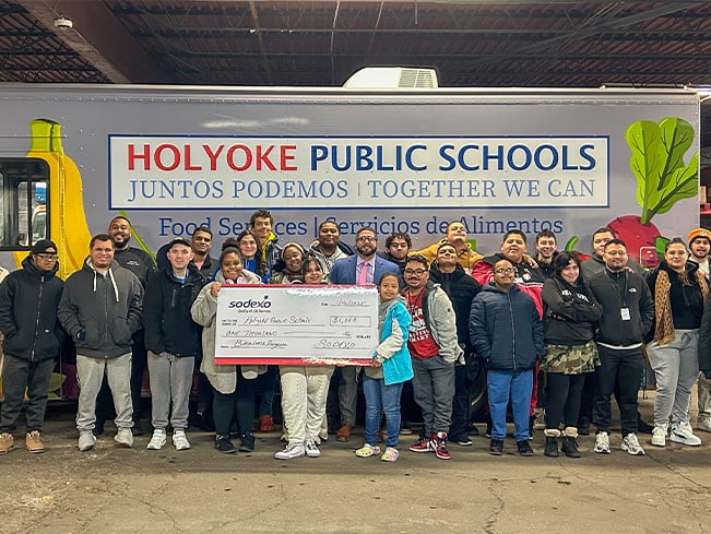 Group of students, families, and community members posing in front of a colorful school food services truck while holding a donation check, highlighting food access programs, community support, and school partnerships.