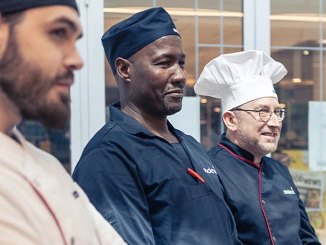 A man wearing a blue chef's coat and hat, smiling, stands in a kitchen setting.