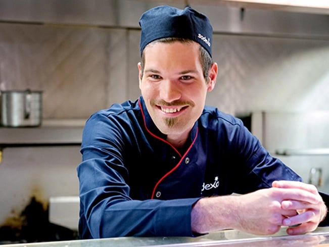 A man wearing a blue chef's coat and hat, smiling, stands in a kitchen setting.