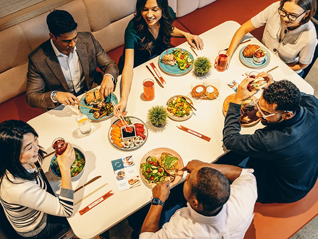 Top view of several people seated around a table sharing a variety of dishes including sushi salads and sandwiches with drinks and condiments in a modern dining setting.