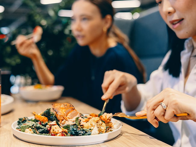 Two people sitting at a table eating a plated meal with salmon and greens, using utensils while another dish and drink are visible in a softly lit indoor dining setting.