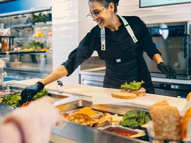 Food service worker wearing an apron and gloves preparing a sandwich at a counter with fresh ingredients including greens sliced vegetables and bread in a bright modern kitchen setting.
