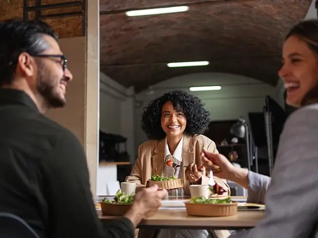 Three people sit around a table indoors, eating salads from bowls in a restaurant or café with an arched ceiling and modern lighting.