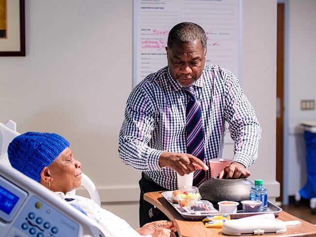 A man in a blue shirt and tie assists a woman in a hospital bed, providing support and care.