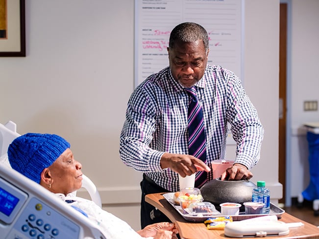 Hospital staff member serves a meal tray to a patient resting in a hospital bed.