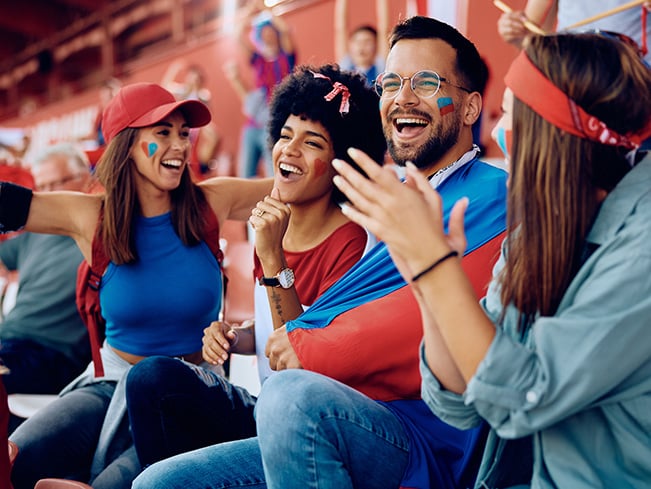 Group of fans in stadium seats wearing team colors, clapping and raising hands, one wrapped in a flag.