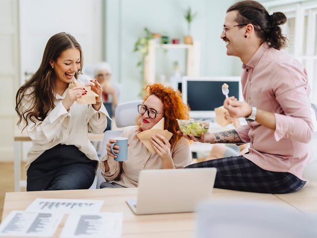 Colleagues share lunch and snacks around a desk in an open office, with a laptop and papers nearby.