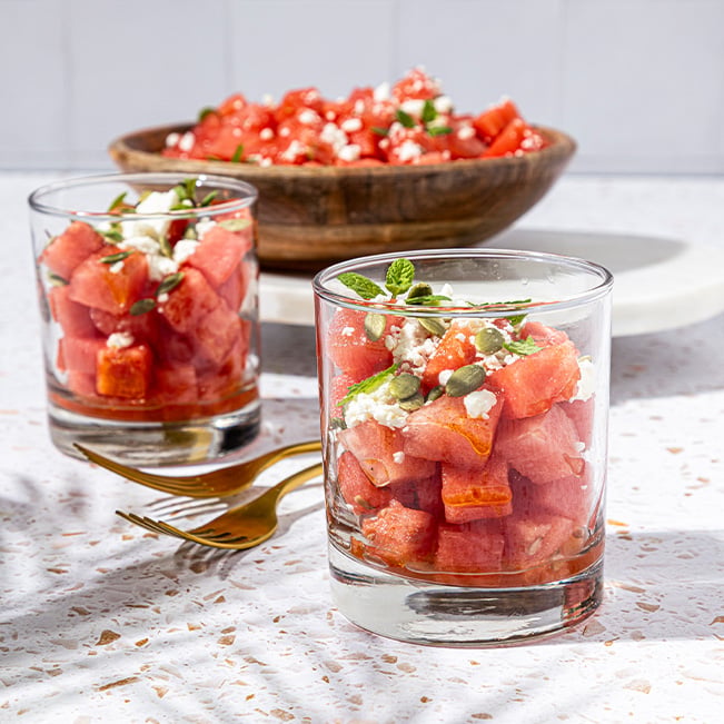 Two glass cups filled with watermelon salad topped with crumbled cheese mint leaves and pumpkin seeds, with a large serving bowl in the background on a bright tabletop.