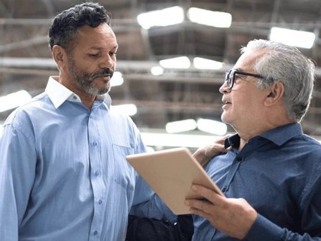 Two men standing side by side in a factory, wearing safety gear and observing machinery in the background.