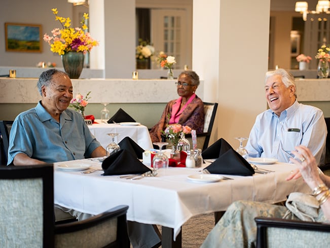 Older adults gathered around elegantly set dining tables in a senior living community restaurant, highlighting social dining, resident engagement, hospitality, and meaningful shared experiences.