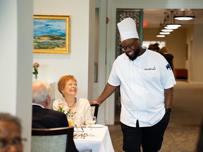 Chef in a senior living dining room speaking with residents at a table, highlighting personalized hospitality, resident engagement, dining satisfaction, and community-centered service experiences.