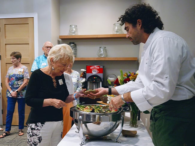 Chef serving a fresh salad from a buffet station to a resident during a community dining event, highlighting hospitality, personalized service, social engagement, and resident-centered dining experiences.