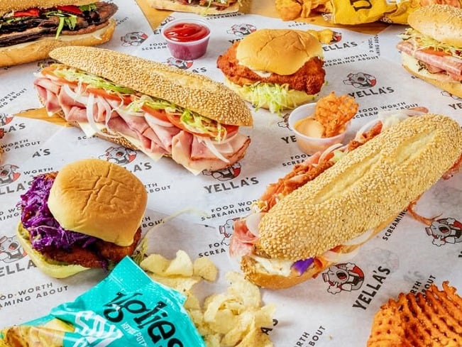 An assortment of Yella’s sandwiches, chips and waffle fries arranged on branded paper.
